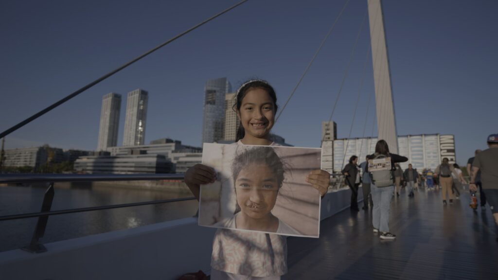 Una niña sonriente sostiene una foto de sí misma antes de su cirugía de fisura labio palatina. Está de pie sobre el Puente de la Mujer en Buenos Aires, con edificios modernos al fondo y personas caminando a su alrededor. La imagen simboliza el impacto transformador de Smile Train en la vida de los niños con fisura labio palatina.